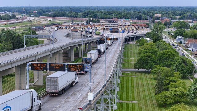 Port Huron, Michigan, United States - 27 July 2026: Aerial view of the Blue Water Bridge Toll Booth with trucks queuing in lanes under electronic signs, surrounded by highway infrastructure.