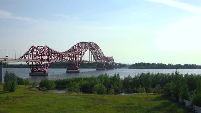 Red steel arch truss bridge spanning wide river with bright sun glare, reflective rippling water, grassy bank, dense trees, and open panoramic daylight landscape