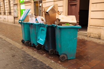 Fototapeta premium Close up of overcrowded blue and green plastic waste containers stacked with trash and discarded cardboard on a sidewalk next to a building.