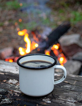 Enamel camping cup with steaming hot coffee resting on weathered log outdoors, glowing campfire nearby, warm orange flames illuminating the rustic wood, cozy woodland setting, soft blurred forest