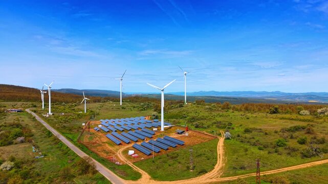 Aerial view of solar farm surrounded by wind turbines. Unpaved dirt paths cross green grassy terrain beneath a bright blue open sky.