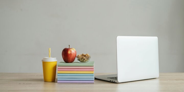 Laptop and stack of colorful books with an apple and snack on a wooden desk