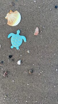 Top view of decorative sea turtle and seashell figures on wet beach sand, as sea wave foam washes in from the side leaving ample copy space for advertising.