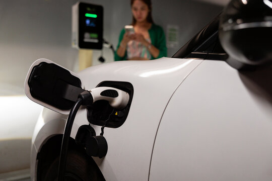 Woman charges electric car, checks mobile phone.