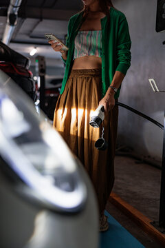Woman holds EV charger and mobile phone in parking garage.