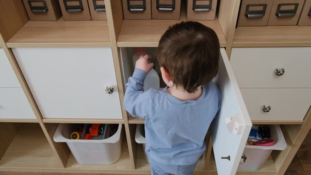 Toddler baby plays with household chemicals and detergent bottle from the closet. Child boy with cleaners products in home living room. Kid about two years old (age one year nine months)