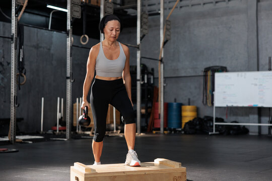 Mature woman performing kettlebell box step-ups in the gym.