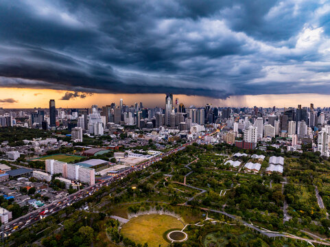 Bangkok, Thailand - 20 June 2024: Aerial view of the Bangkok skyline and Benjakitti Park in Khlong Toei under a dramatic dark storm cloud at sunset, featuring tall skyscrapers and lush green parkland.