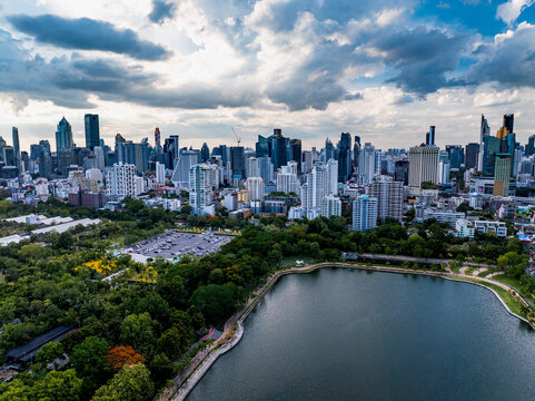Bangkok, Thailand - 20 June 2024: Aerial view of Benjakitti Park with its lake and lush greenery set against the modern high-rise skyline of Bangkok under a cloudy sky.