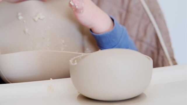 Child eating oatmeal with spoon at dining table. Messy toddler learning to self feed. Healthy baby breakfast, nutrition and growth development. Concept of messy meal time at home.