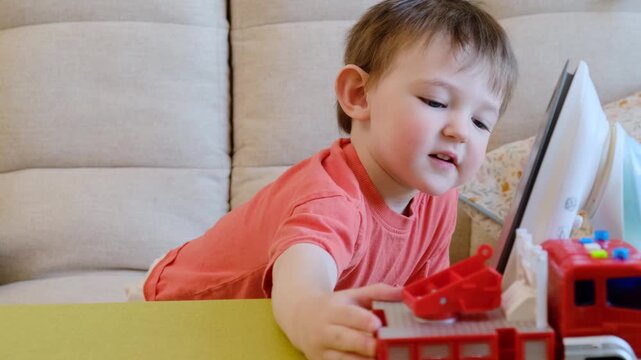 Child boy sitting near hot iron and playing with toy fire truck on table. Household danger and safety education for toddler. Risk of burn injury and hazardous home environment scene.