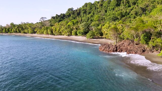 Drone flying along the scenic tropical coastline of San Josecito beach, Osa Peninsula, Costa Rica. Clear turquoise ocean, gentle waves breaking on a rocky outcrop and sandy shore.