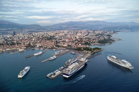An aerial view highlights luxury yachts and cruise ships anchored in the sapphire harbor of Split, framed by the coastal mountains and historic red roofs of Dalmatia, Croatia.