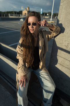 High-angle shot of a stylish young woman in a beige trench coat and sunglasses posing outdoors in golden hour sunset light. Pretty autumn beauty girl
