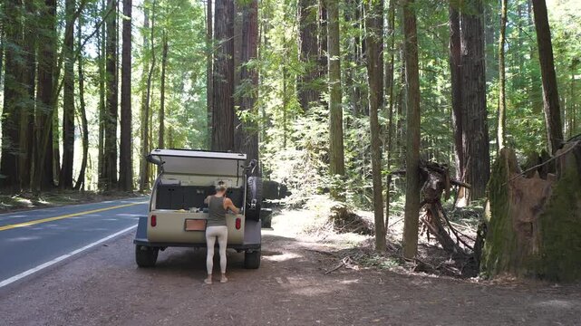 A woman takes a lunch break by her teardrop camper trailer in Navarro River Redwood State Park, enjoying views of the forest and river.