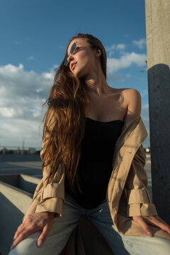 Chic woman with long wavy hair and sunglasses posing in a loose beige trench coat on a concrete rooftop, looking towards the cloudy sky on a sunny day.