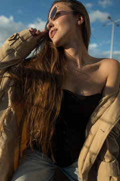Close-up portrait of a gorgeous young woman in sunglasses touching her hair, wearing a beige trench coat and black top, posing outdoors under a bright blue sky.