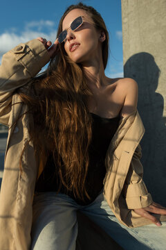 Confident young woman with long wavy hair wearing sunglasses and a beige trench coat posing against a blue sky on a sunny day in an urban setting. Autumn beauty lady