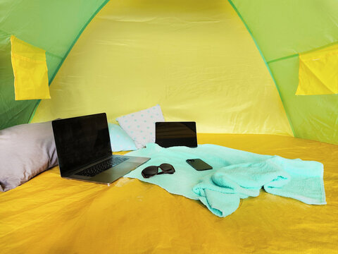 Laptop, smartphone, sunglasses and pillows inside a camping tent with colorful fabric interior. Cozy travel setup for remote work, digital nomad lifestyle, camping vacation and outdoor leisure.
