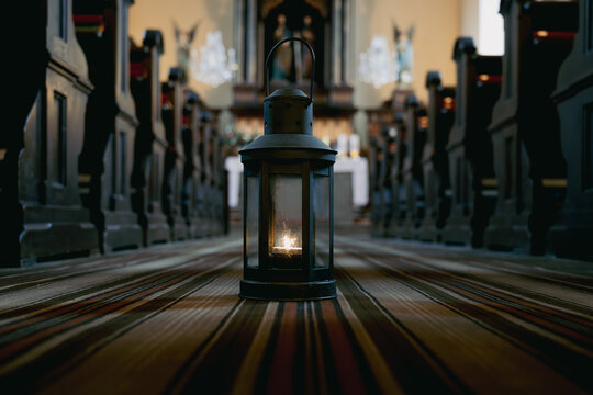 A lantern stands in the center of an empty church, casting a warm light. The wooden pews are lined up on both sides, creating a pathway. The setting shows a peaceful moment in time.