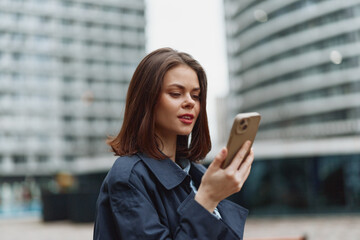Fototapeta premium Young woman with straight brown hair wearing a dark coat looks at her smartphone with a slight smile in an urban environment with modern buildings in the background.