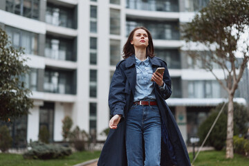 Fototapeta premium Woman outdoor fashion urban denim coat smartphone confidence posing street, looking up with a calm gaze in a sunlit moment, modern architecture in the background