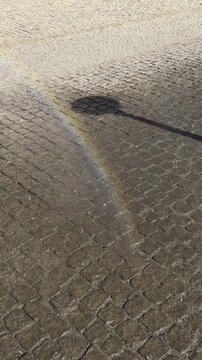 Fine water spray from a public street fountain creating a colorful rainbow above wet and dry stone pavement, urban summer scene with sunlight, water droplets and colorful refle
