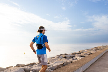 Person walks away along a rocky ocean path. They wear a blue shirt and a hat. The sky shows soft clouds and sunlight. Their posture suggests calm reflection. This image suits health or travel themes © VICTOR