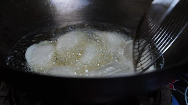 Close up of food frying in hot oil in a dark pan with bubbles and a metal skimmer, showing cooking in the kitchen
