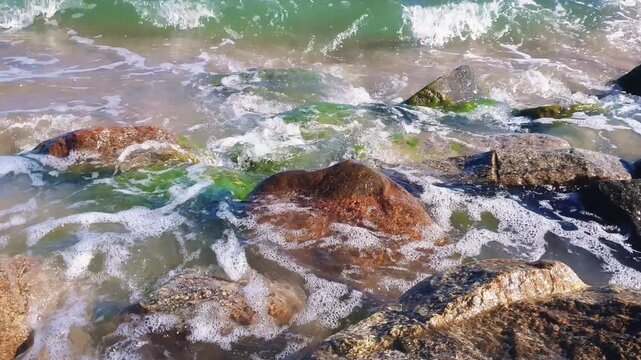 Close-up of foamy coastal surf with azure stormy sea waves crashing on wet rocks and seaweed covered stones on rocky shore of Baltic sea at sunny day. Seaside water surface pattern natural background.