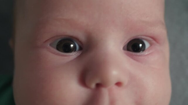 Very close frontal view of infant face and eyes showing slight movement, pursed lips, moist lower lip, small chin creases, soft skin texture, gentle green collar, calm indoor setting