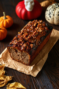 A nut-topped pumpkin bread loaf on brown parchment paper surrounded by small pumpkins, a skull and autumn leaves.