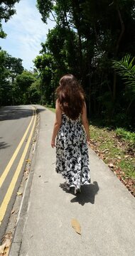 Follow shot of tourist woman in long patterned dress and ballerinas walking on sidewalk along road in tropical park, enjoying sunny weather and peaceful nature of Singapore with no one else around.