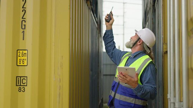 A senior logistics worker uses a digital tablet for inventory management, standing between shipping containers at a cargo port to streamline supply chain operations.