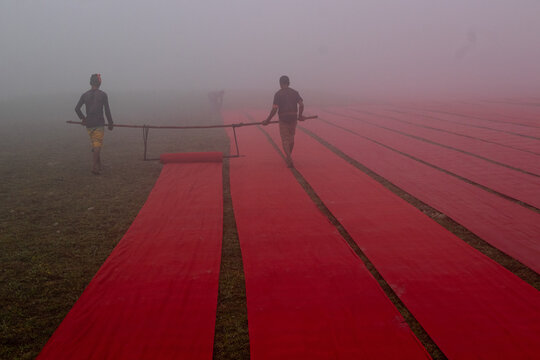 Narsingdi, Dhaka Division, Bangladesh - 22 January 2025: Aerial view of workers carrying a long roll of vibrant red fabric across a field during a foggy morning.