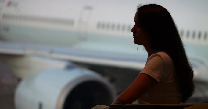 Pensive woman traveler sit against large window, slow going time while waiting for boarding gate to open. Almost silhouetted view of passenger against large airplane parked at gate blurred on back