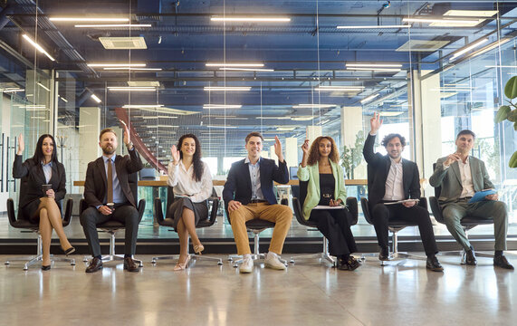 People in business team meeting raise hands. Professionals sit aligned in a glass office, joining a workshop by voting and showing enthusiasm. Theme of teamwork and active participation.