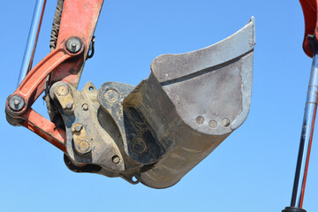A close up of a heavy duty metal ditching bucket attached to an orange excavator hydraulic arm under a clear blue sky. © nahhan