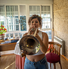 Young woman plays trumpet indoors beside large bright windows, framed by warm daylight, textured wall, wooden chair, and casual home setting, showing focused practice and musical performance © carlos