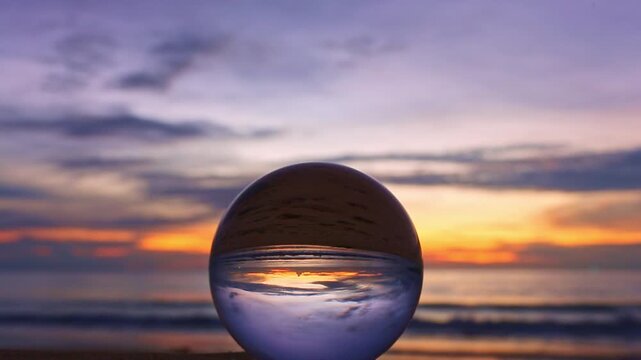 A glass sphere placed on the beach reflects an inverted view of a vibrant sunset over the ocean, creating a striking contrast between the warm golden sky and the deep blue clouds within the orb