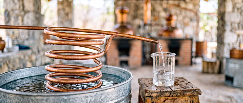 Copper coil moonshine still pouring clear spirit into glass beaker in rustic distillery