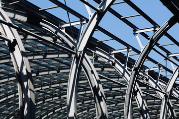 Fototapeta premium Steel roof structure under sky. Curved metal beams above glass. Gray columns under arched framework. Repeating steel sections across roof. Blue sky in background. Modern steel architecture detail.