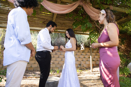 Couple exchanging rings under pergola draped pink fabric in garden, bride wearing white lace gown