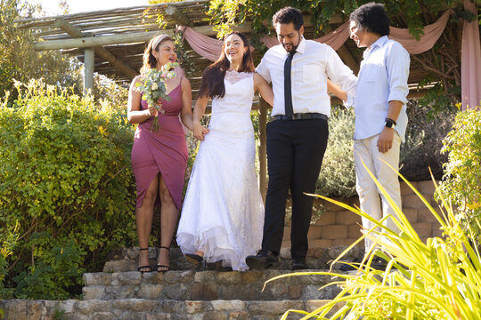 Diverse bridal party descending pergola steps draped pink, bride in white lace gown holding bouquet