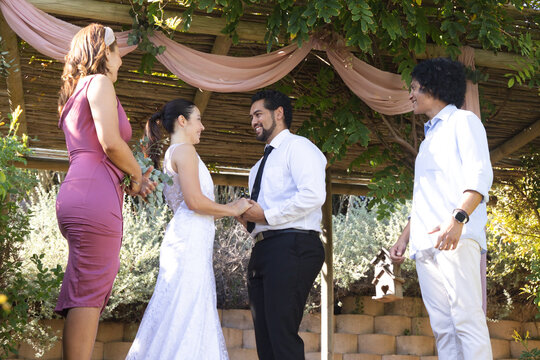 Couple exchanging vows under pergola draped pink fabric, bride in white lace, groom in black tie