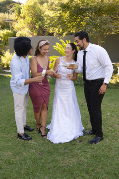 Diverse group toasting on garden lawn, bride in white gown, groom pouring champagne into flutes