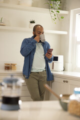 African American man leaning in bright kitchen wearing denim shirt holding grey mug checking phone © wavebreak3