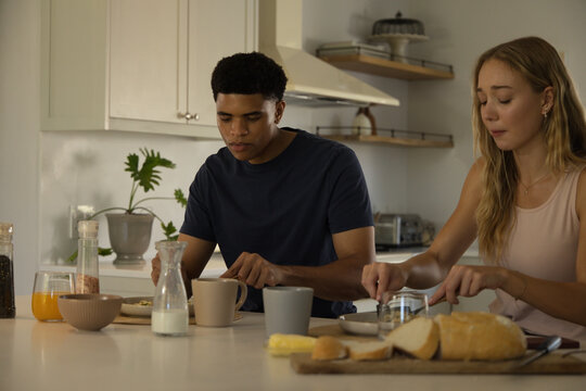 Diverse couple preparing breakfast at kitchen island, spreading butter on sliced bread