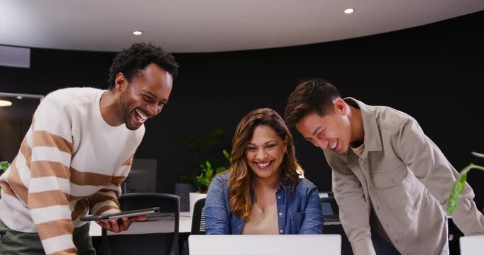 Diverse coworkers leaning in and laughing while scanning laptop after tablet alert at office