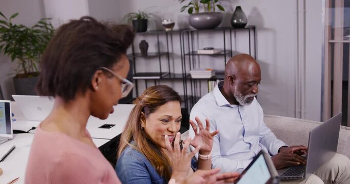 Diverse coworkers meeting in lounge, standing woman turning tablet senior man shifting from laptop